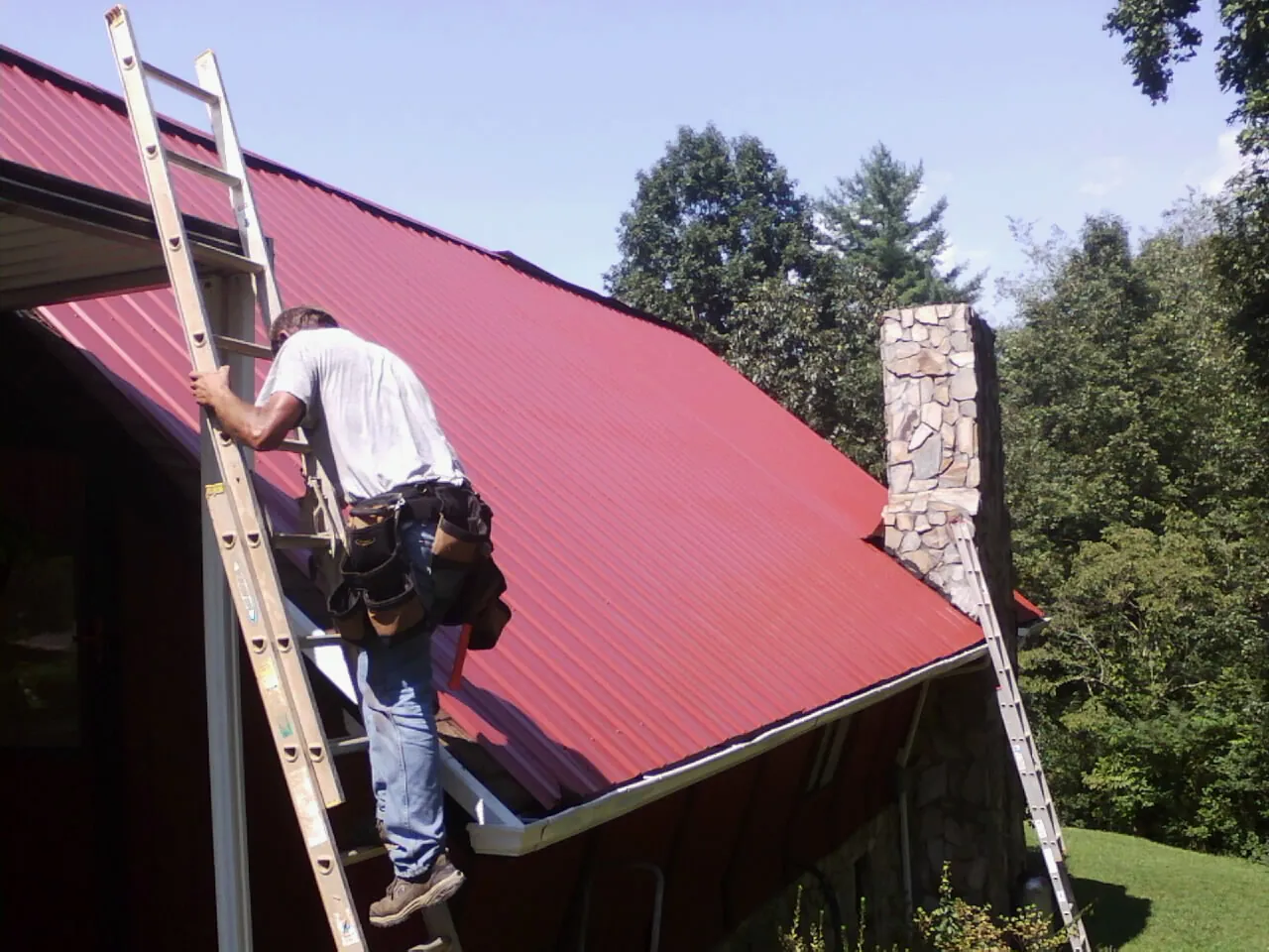 Roofer on a ladder beside a red metal roof and stone chimney — Soffit & Fascia Repair by Roofing Brothers in Foxburg